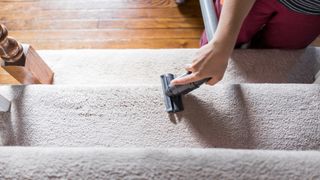 A close up of a young woman's hand pictured holding a vacuum cleaner attachment to carpeted stairs