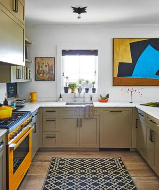 kitchen with white walls, taupe cabinets, orange range, bold artwork, black patterned rug and wooden floor