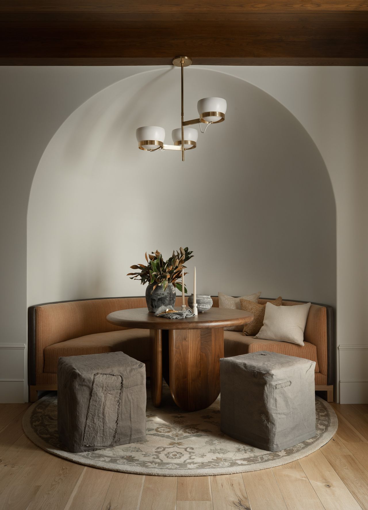 Curved arched inset dining area with curved banquette seating a round dining table, a round rug, two square poufs and foliage on the table with a ceiling pendant hanging over the table