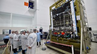 A technician in the cleanroom explains details about the Eutelsat Quantum satellite, to a crowd small crowd as they all stand in a cleanroom