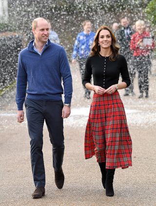 Kate Middleton wearing a red plaid skirt and black sweater with fake snow falling behind her while walking next to Prince William wearing a blue sweater and gray pants