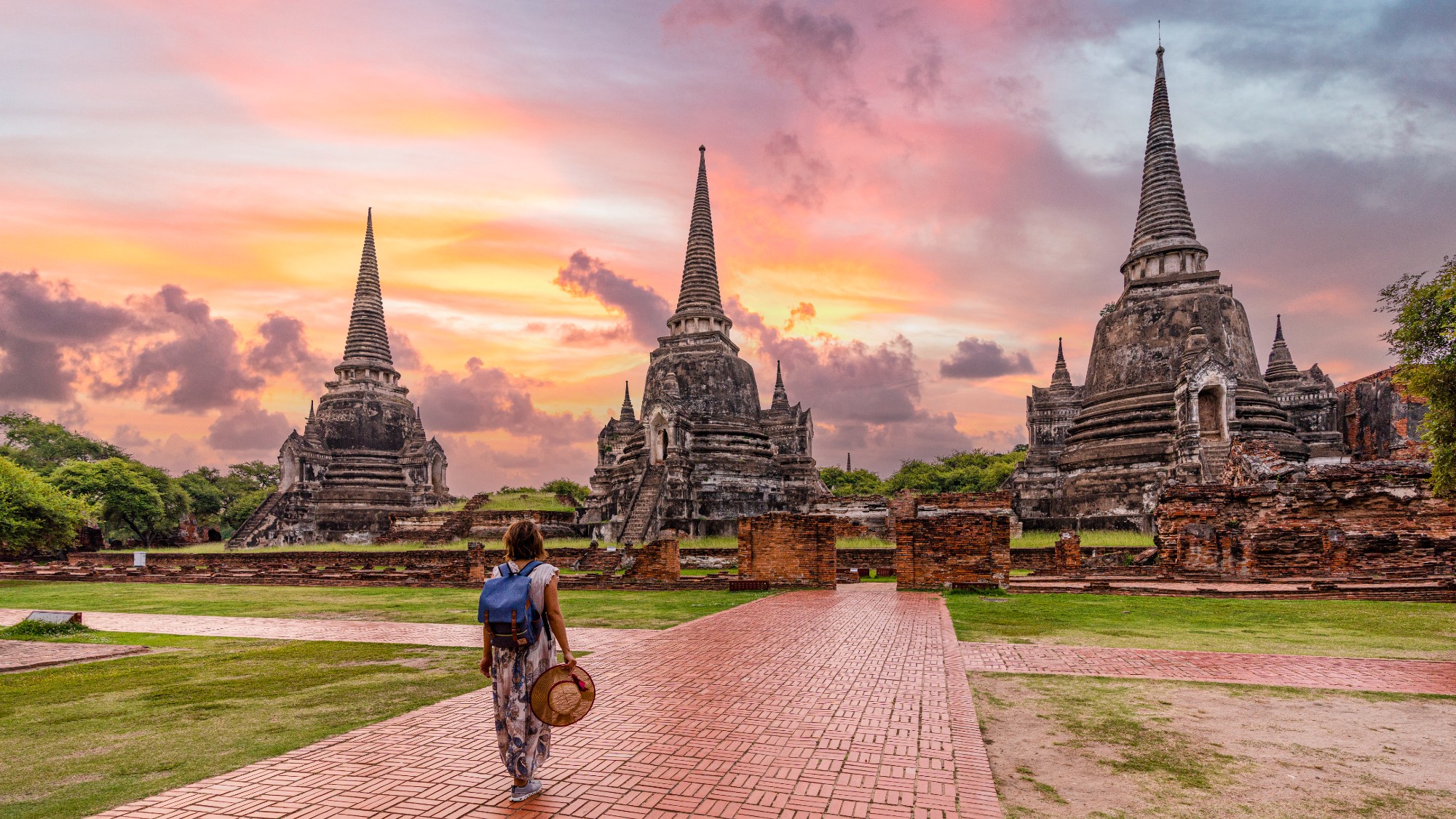 A woman holding a hat in her head stands in front of Wat Phra Si Sanphet Buddhist temple in Thailand