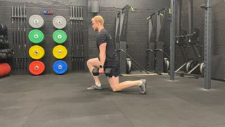 Fitness writer Harry Bullmore performs a dumbbell lunge in a gym. We can see colorful weight plates and fitness equipment behind him. He faces away from the camera, with his left knee bent and grazing the floor and his right knee bent in front of him.