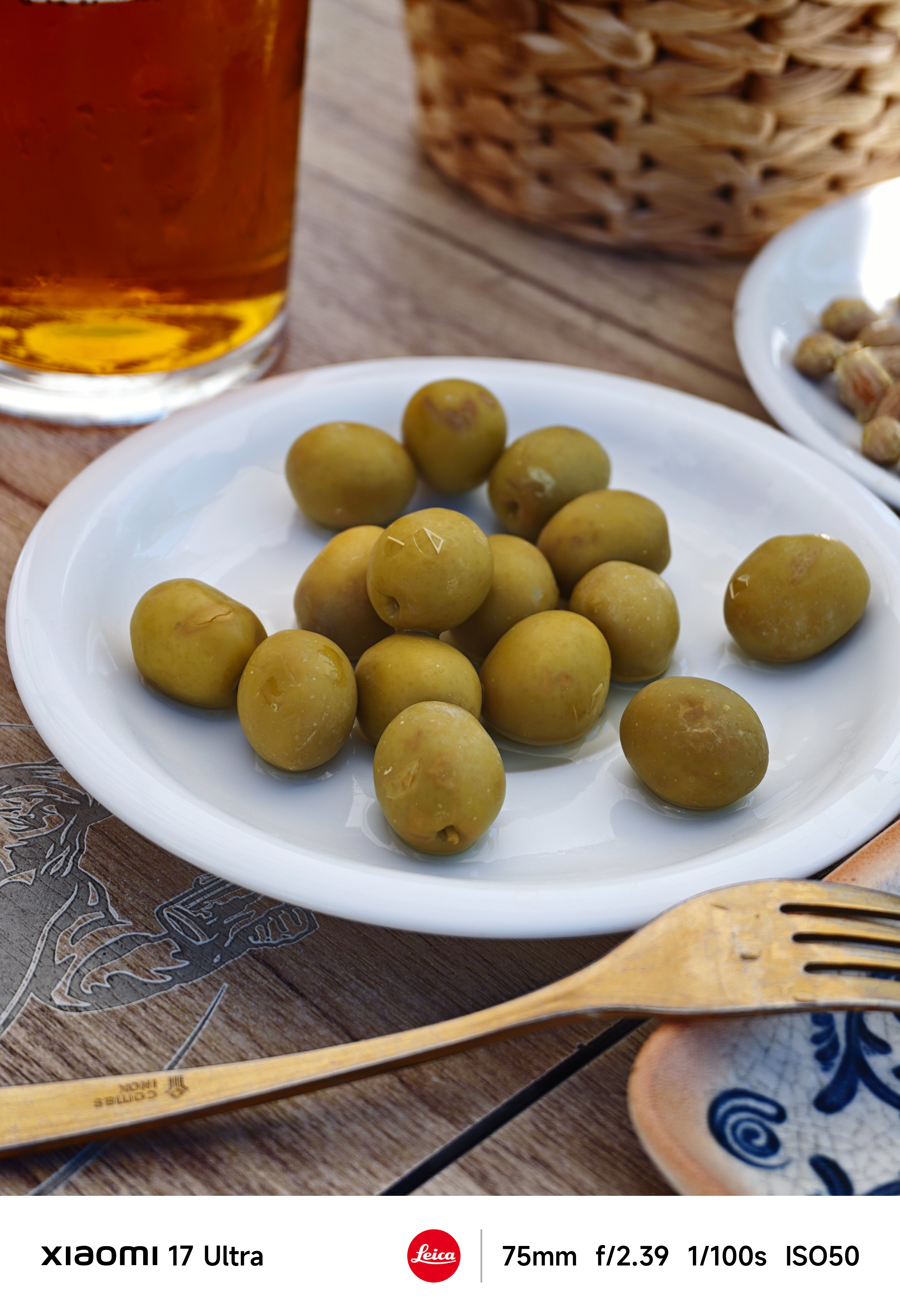 White plate of green olives on a wooden table with a glass of beer and small dishes in the background.