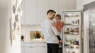 Man holding a toddler looking into an open fridge in a modern white kitchen.