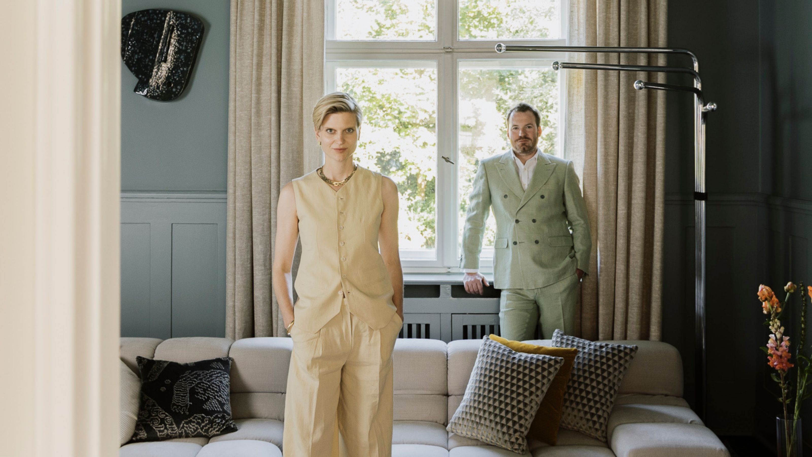 A woman in a two-piece, sleeveless beige casual chic suit, and a man in a double-breasted pale mint one stand in a room painted pale blue surrounded by beige curtains, a modular sofa, and artwork.