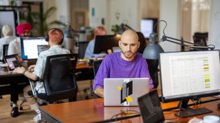 Platform engineering team working in an open plan office space with male software developer in purple shirt pictured at desk in foreground.