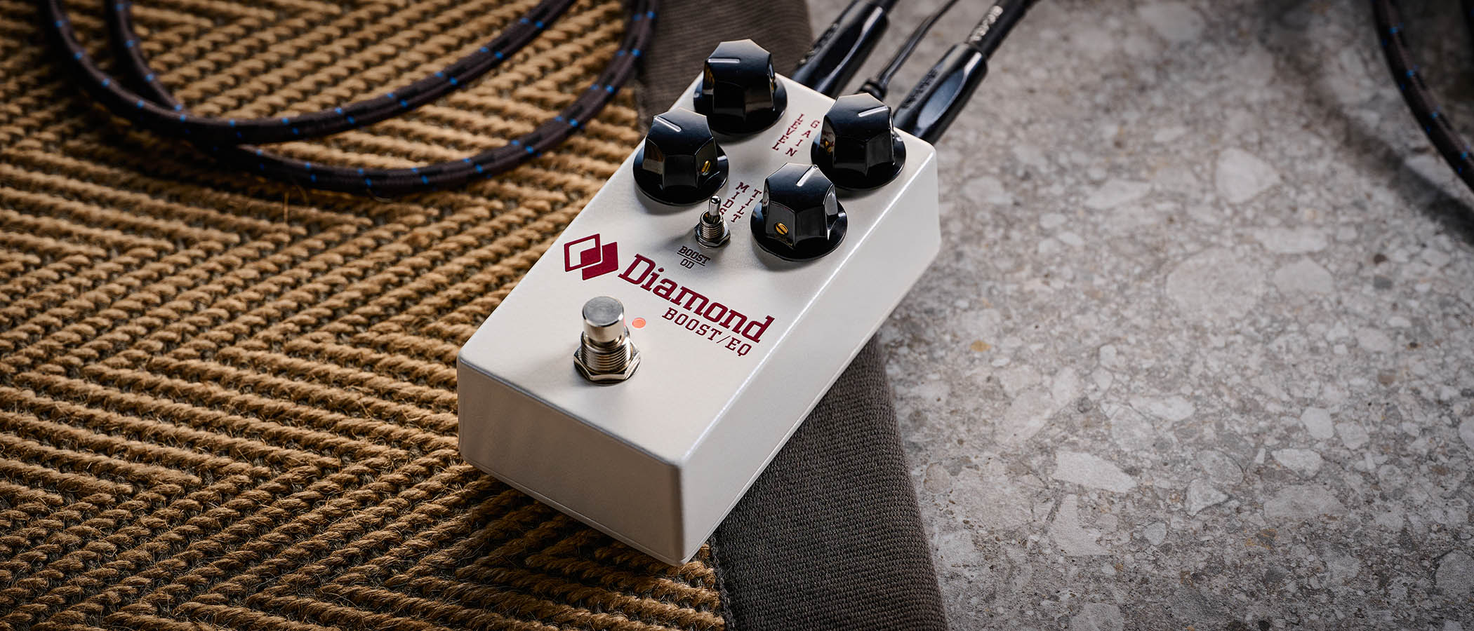 The Diamond Boost/EQ is a compact four-knob pedal in a white enclosure with black knobs and a toggle switch, and is photographed here in close-up against a patterned rug.