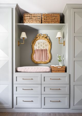 gray and pink nursery with custom wardrobe and drawer space, gold wall lights, gold ornate mirror, shelving with rattan boxes, changing area