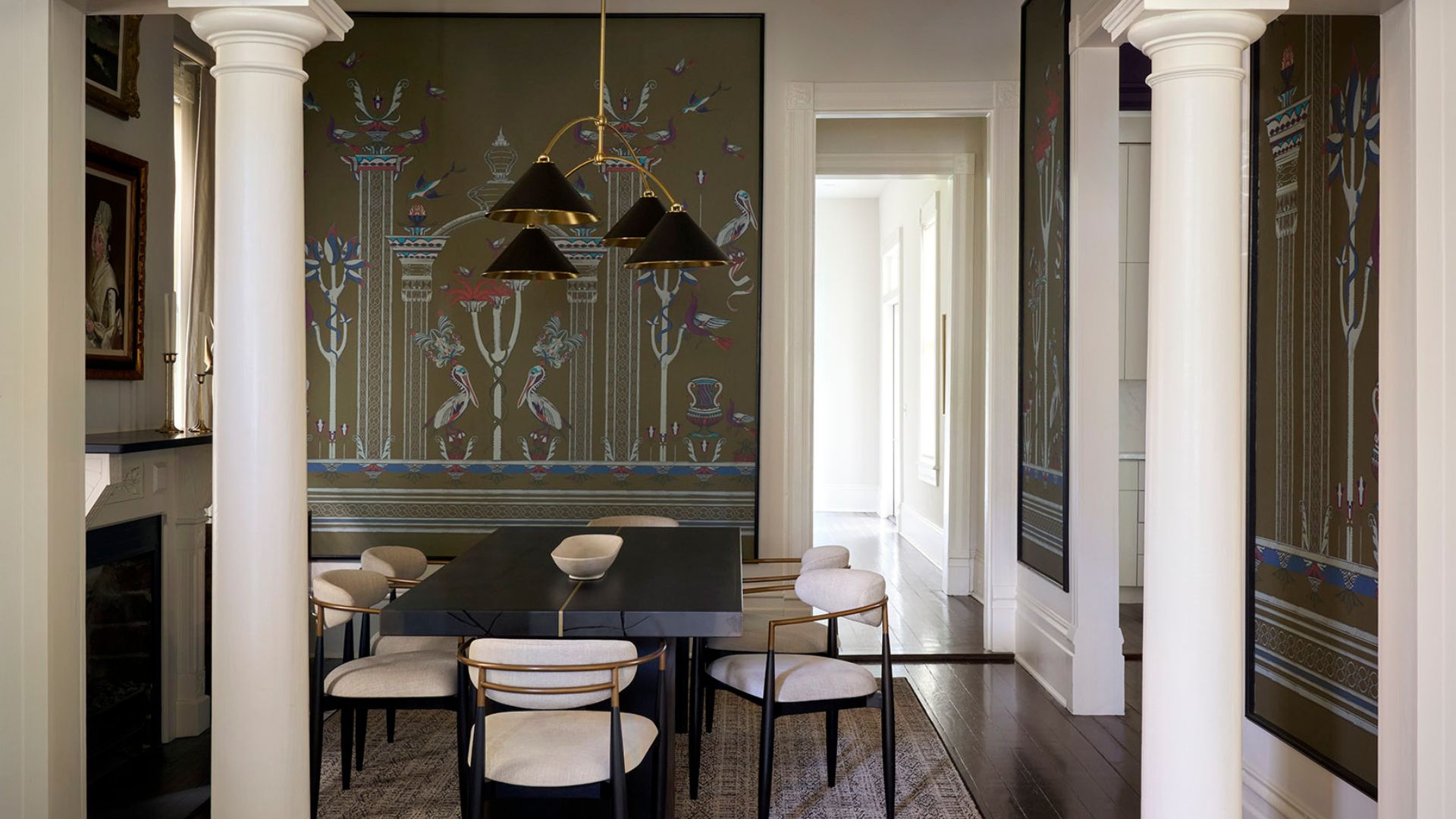 A white dining room with framed wallpapers, a black table, white armchairs with gold accents, a pendant lamp and a rug