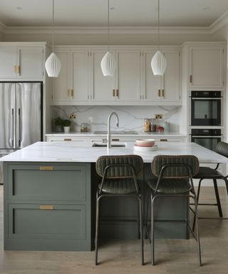 A kitchen with white cabinets with gold handles, a kitchen island with a marble worktop, sage green cabinets with gold handles and two dark green stools underneath it, and three white pendant lights on the ceiling