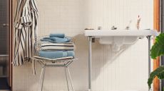 Retro-style bathroom featuring white ceramic tiles and a mid-century inspired chrome chair, which is holding a stack of fluffy bath towels. A coordinating robe hangs alongside.