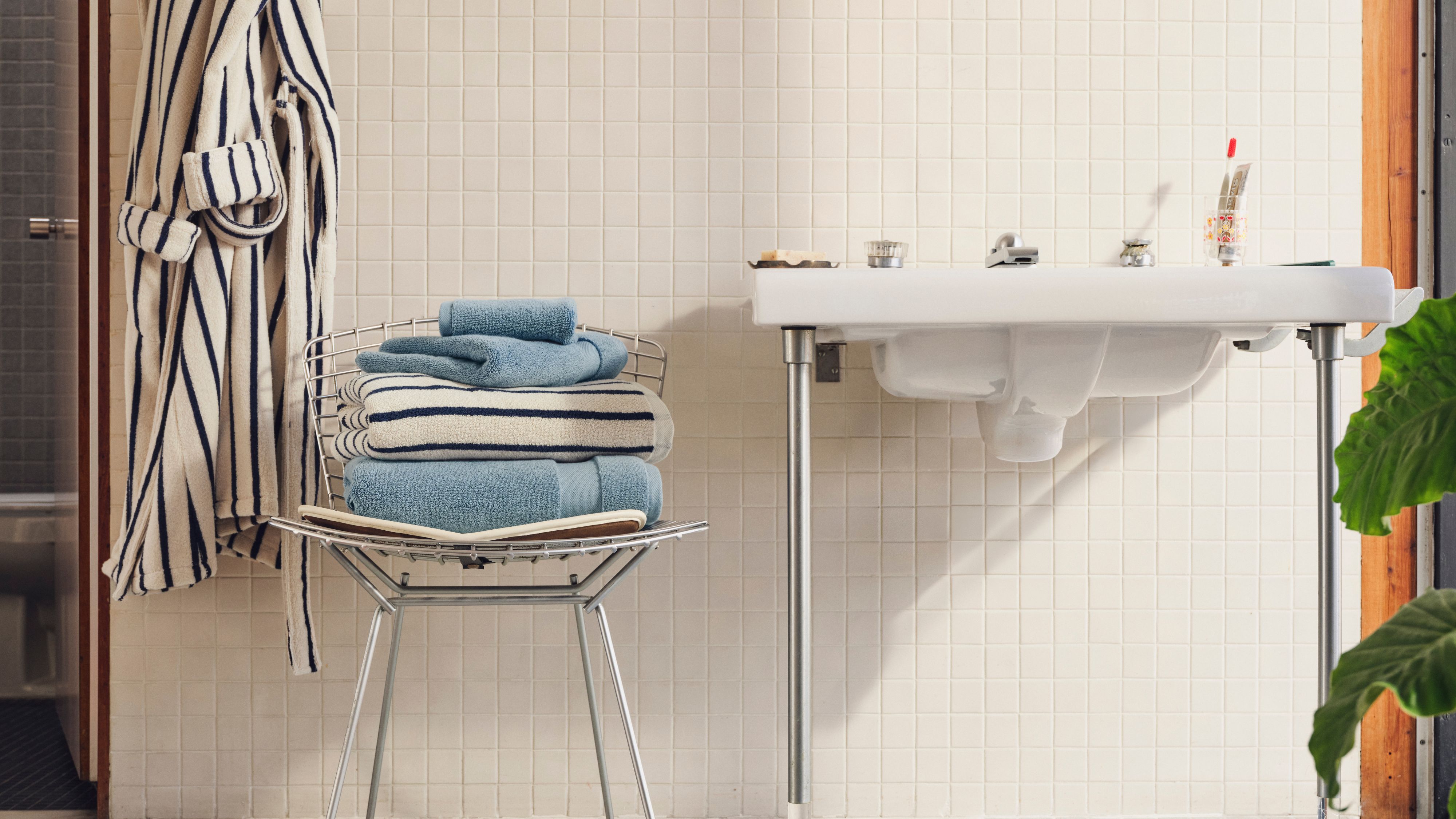 Retro-style bathroom featuring white ceramic tiles and a mid-century inspired chrome chair, which is holding a stack of fluffy bath towels. A coordinating robe hangs alongside.