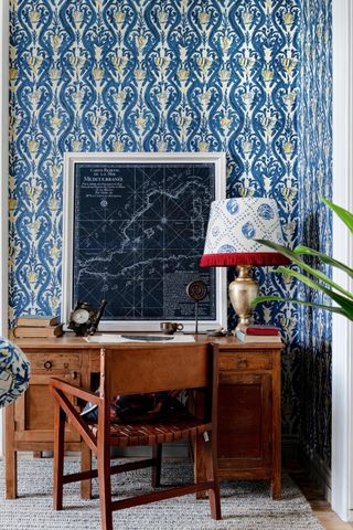 home office with vintage desk and chair, bold blue and yellow damask style wallpaper, gold based table lamp with blue and white shade with red fringing, large map print on desk