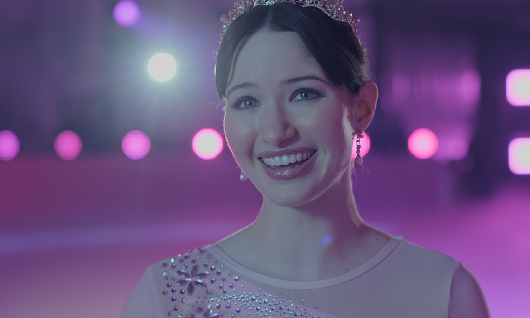 Madelyn Keys as Adriana Russo wearing a tiara and pink ice skating dress smiling while on an ice rink draped in pink lighting in a still from finding her edge