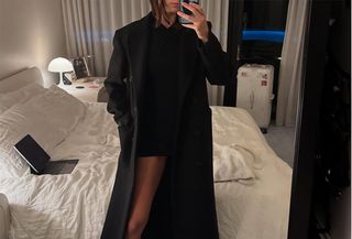 Devon Lee Carlson taking a mirror selfie wearing a black sweater with a long black coat and colorful pointed toe heels.