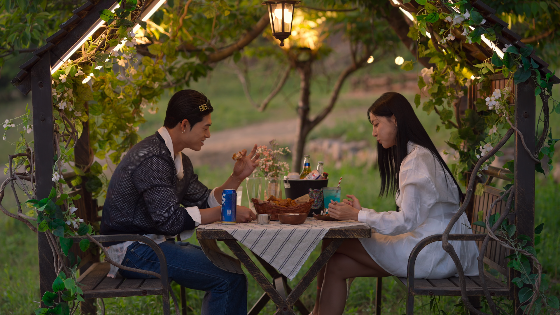 A man (Song Seungil) and a woman (Kim Min-gee) share a meal while sitting in a wooden outdoor table with attached booths and a lit roof. The man wears a beanie, a gray sweater, and jeans, while the woman wears a gauzy white dress. A still from the Korean reality show 'Single's Inferno' season 5.
