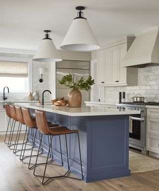 Kitchen lighting ideas with oversized white lampshades hanging over an island with a marble coountertop, blue cabinetry and brown leather bar stools