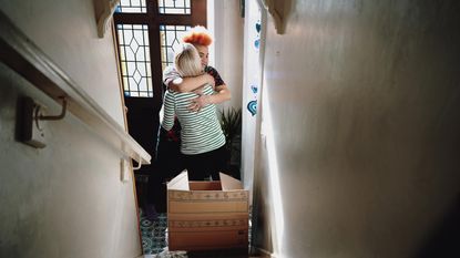 Two women hug at the bottom of a staircase with a moving box sitting on the steps.