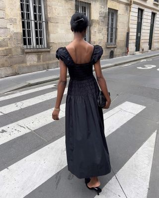 wanita mengenakan maxi dress lengan puff Doen hitam dengan sepatu mule runcing memegang kopling hitam melintasi trotoar jalan