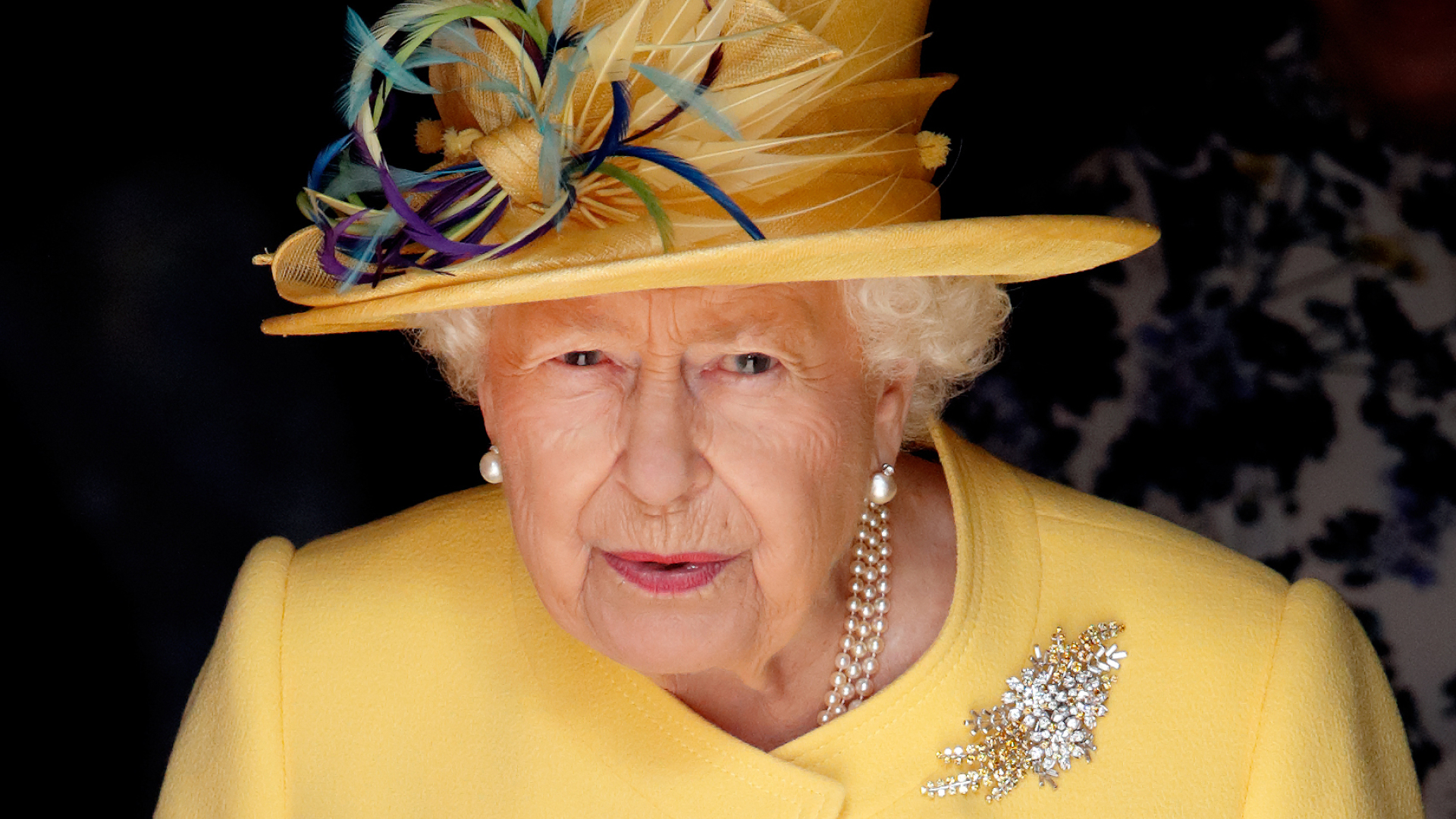 Queen Elizabeth wears a bright yellow coat and a matching hat with purple and blue feathers attached