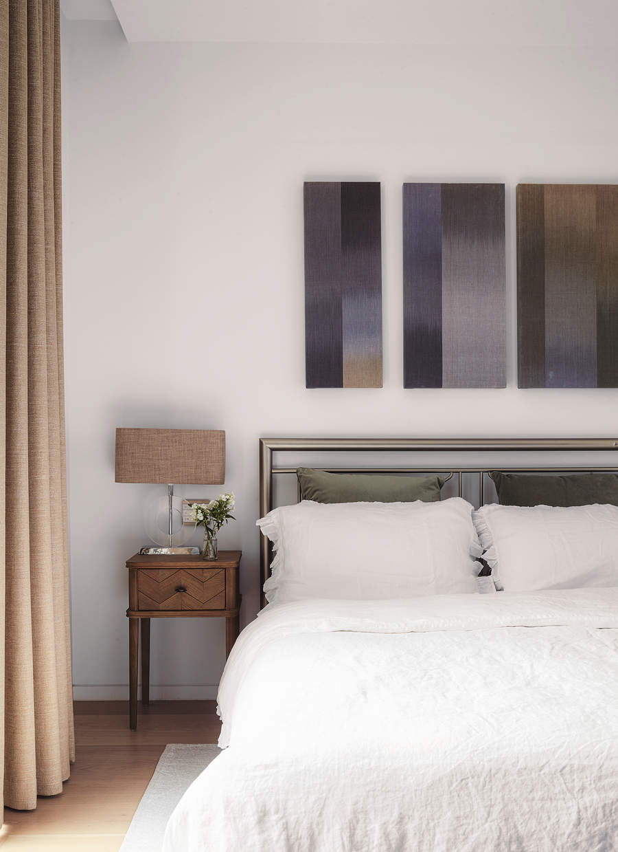 Image of a white bedroom with a bed that has all white bedding except for two velvet, olive green pillows. There is abstract, blue art hanging on the walls and a small wooden side table.