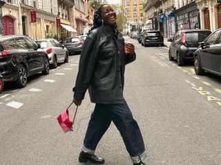 Raïssa Tchoulague on the street in Paris wearing a chic fall outfit, styled with dark blue denim jeans, a longline black leather jacket, loafers, a pink and red handbag, and a black headscarf.