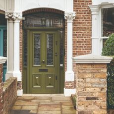 An exposed brick house exterior with a porch and a front door painted olive green with stained glass panels
