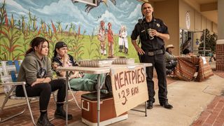 Devery Jacobs as Elora and Paulina Alexis as Willie sit at a table selling meat pies, while Zahn McClarnon as Big is drinking an energy drink