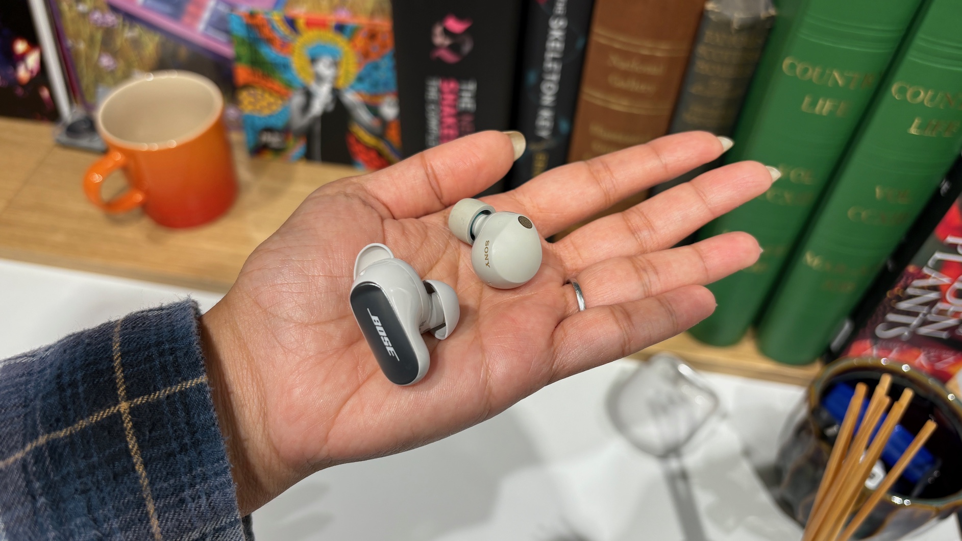 A single earbud of both the Bose QuietComfort Ultra Earbuds (2nd Gen) and Sony WF-1000XM5 held in a hand in front of a shelf full of books.
