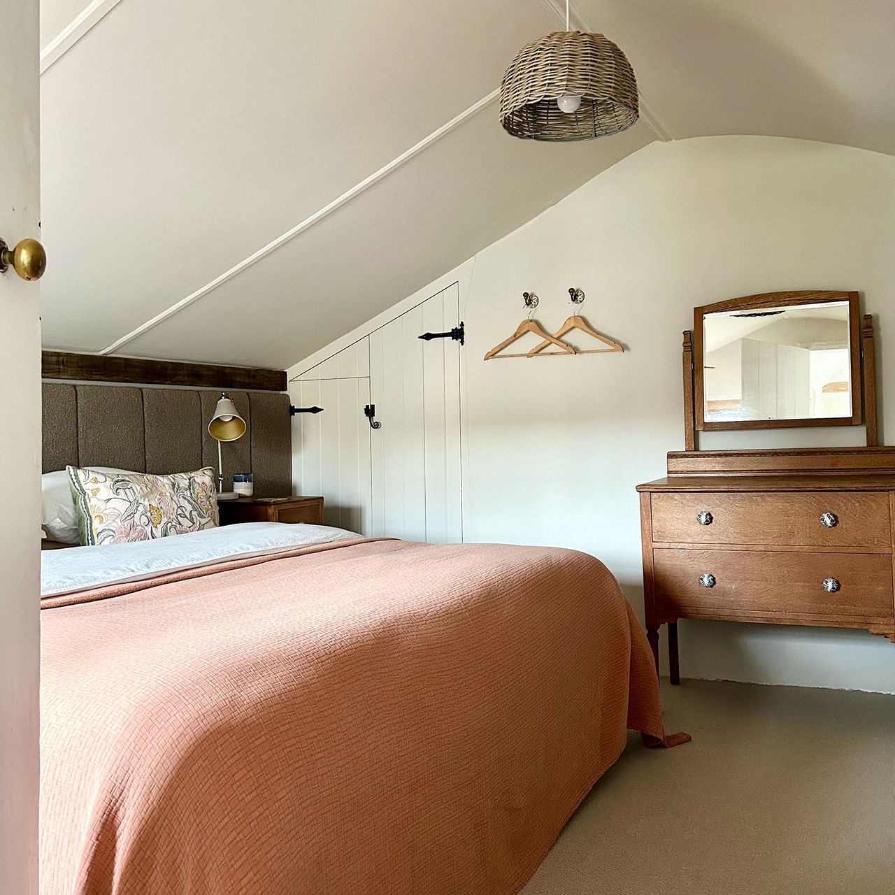 Country bedroom under eaves with wicker lampshade, wooden dressing table and double bed with coral pink throw