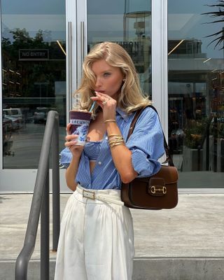 Elsa Hosk wears a chocolate brown Gucci bag with a blue-and-white striped button-down and white trousers with a belt.