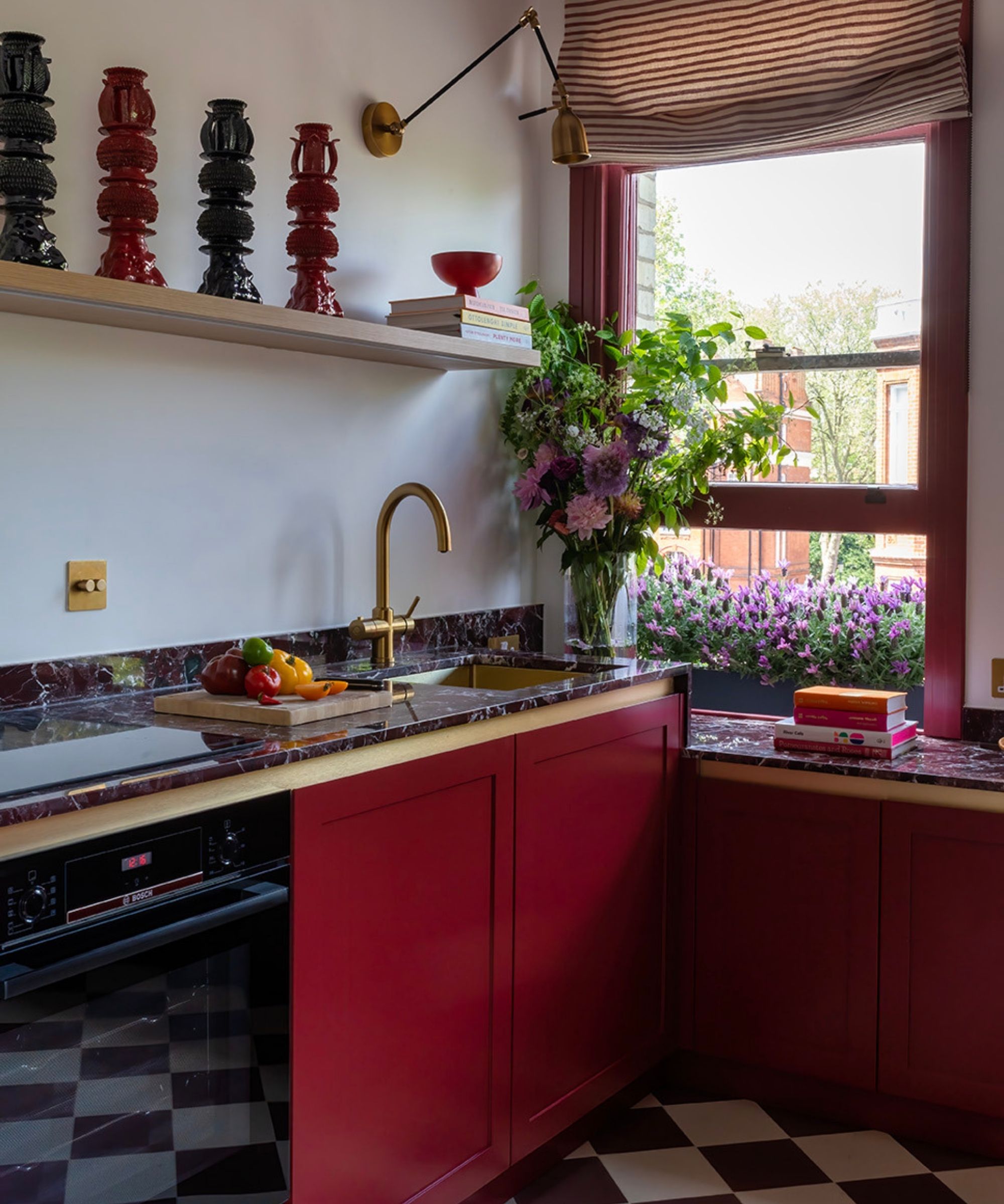 A red kitchen with red cabinets and painted open windows, red marble countertops, gold tap, red and white checkered floors, and a large glass vase filled with flowers.