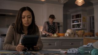 Still from the T.V. show For All Mankind (season 4, episode 1). A young woman with long brown hair is sitting at a kitchen table looking down with an anxious expression at the tablet in her hands. Just on the right of the table you can see a baby blanket and bottle. In the background you can see an older lady cooking in the kitchen.
