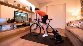 Cyclist riding a bike on a turbo trainer whilst Zwift is displayed on a large TV in front