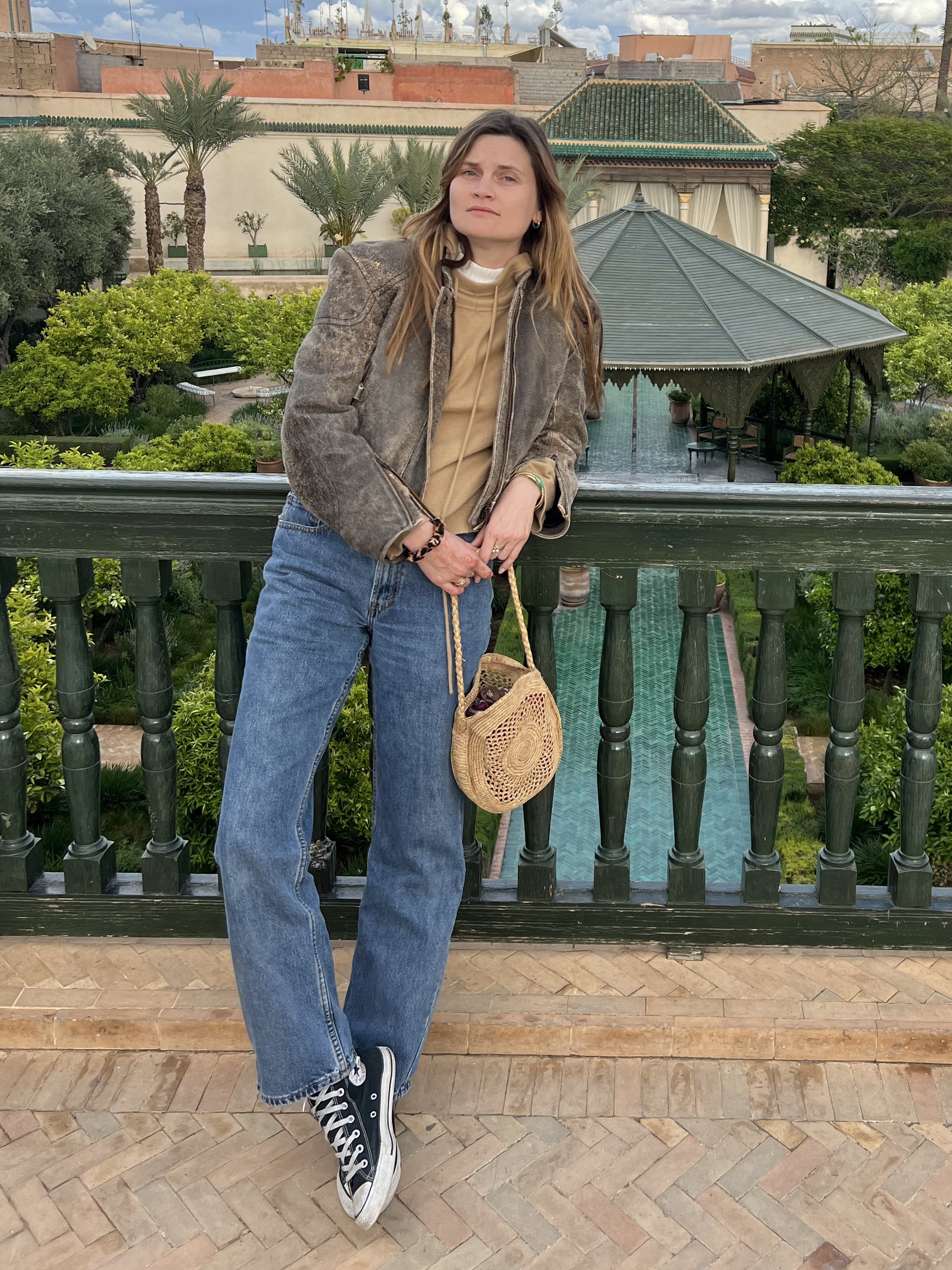 French editor and fashion creative Eugenie Trochu poses against a railing in Morocco wearing a distressed brown leather Acne jacket. a camel sweater, straight-leg jeans, woven bag, and Converse sneakers