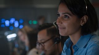 Still from the T.V. show For All Mankind (season 4, episode 1). Close up of a young woman sitting with a row of other people at a console desk. She has shoulder-length dark hair and is smiling. She is wearing a headset and a blue shirt with a collar.