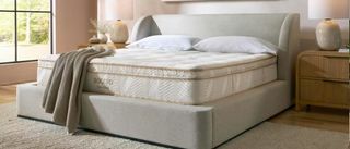 The image shows a cream mattress with gold piping on top of a pebble-colored bed frame, placed in a neutrally decorated bedroom