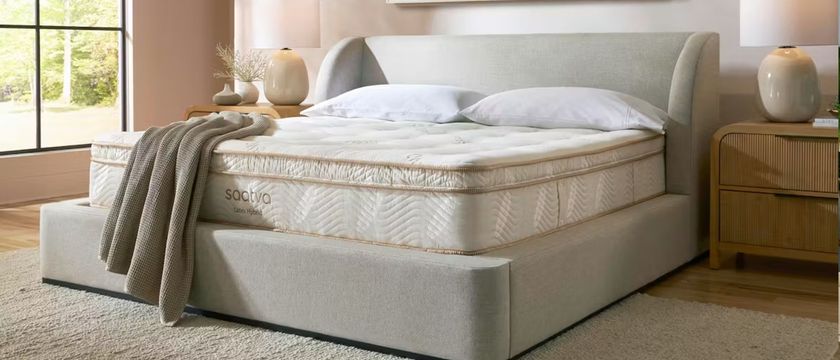 The image shows a cream mattress with gold piping on top of a pebble-colored bed frame, placed in a neutrally decorated bedroom