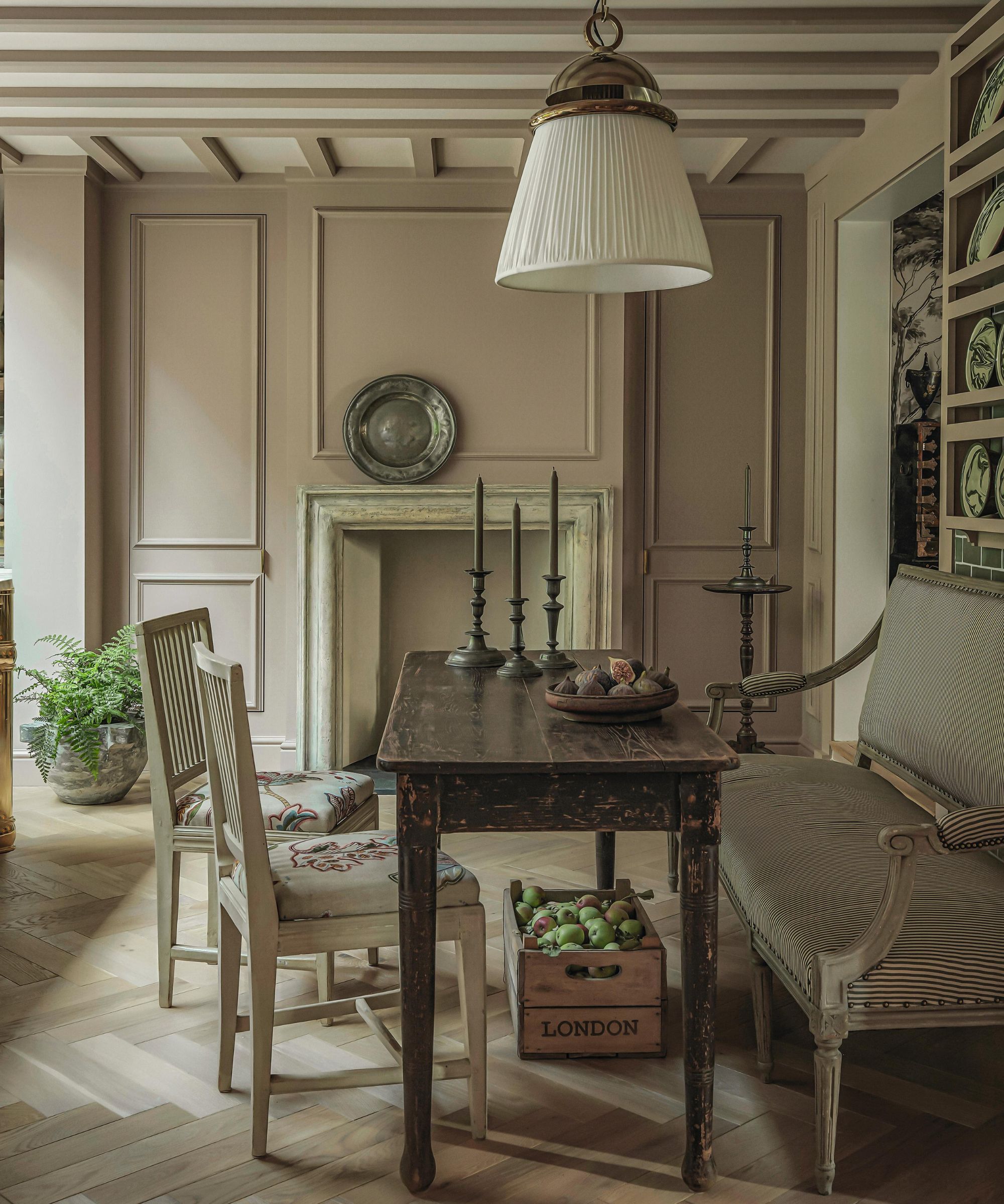a soft pink kitchen dining area with an antique banquette, rustic table and a large pendant light overhead on a beamed ceiling