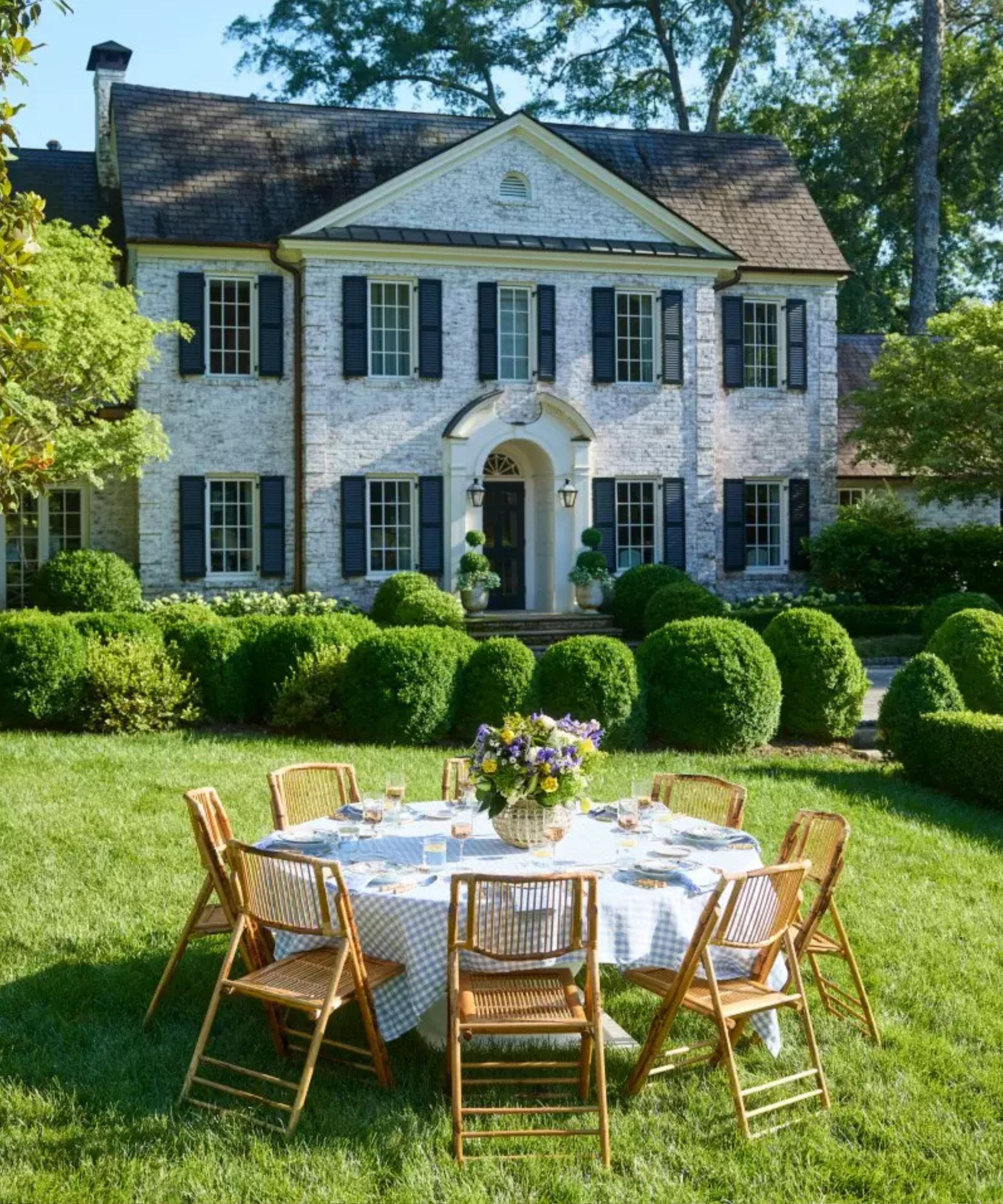large white stone home with a garden lawn, round table with rattan folding dining chairs, blue gingham table cloth laid for dinner