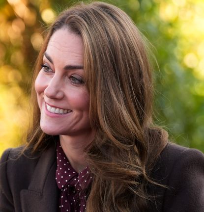 Kate Middleton wearing a brown coat and burgundy polka dot dress smiling with green trees behind her