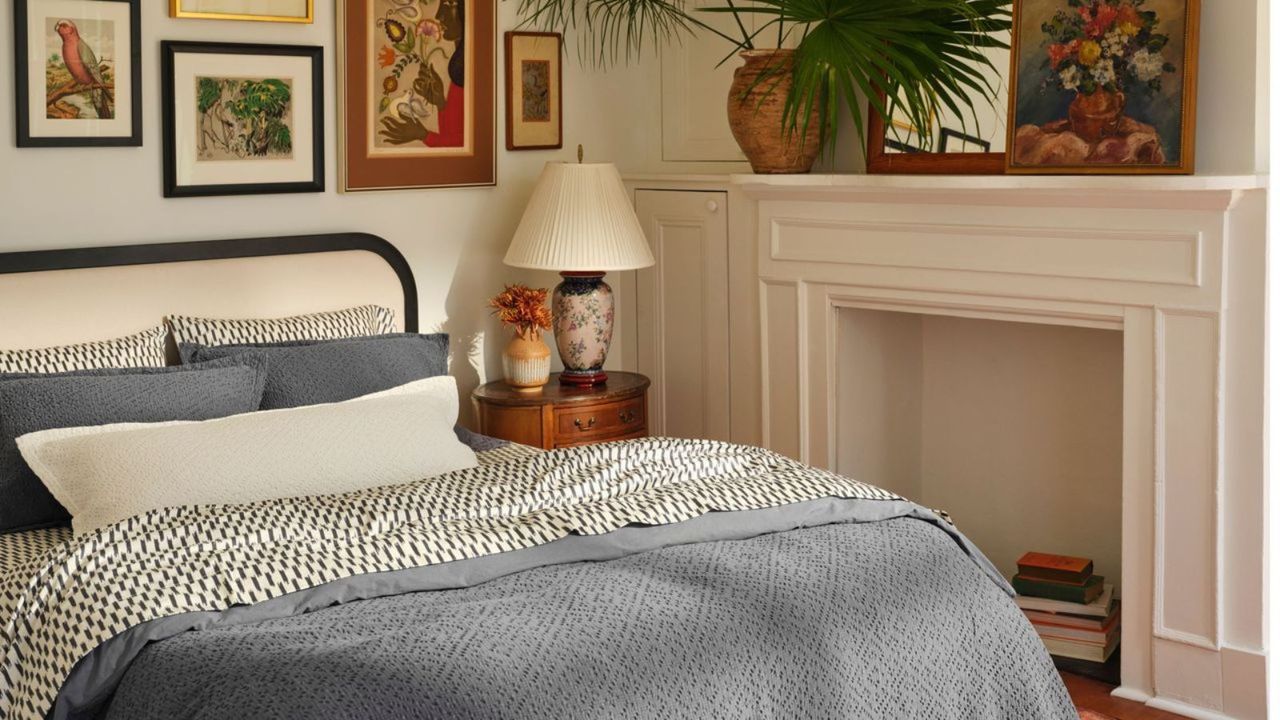 Black and white sheets on a bed with blue shams and blankets; to the right, flowers and a lamp on a dark wood nightstand; a vase, a mirror, and colorful wall art resting on a mantel. 