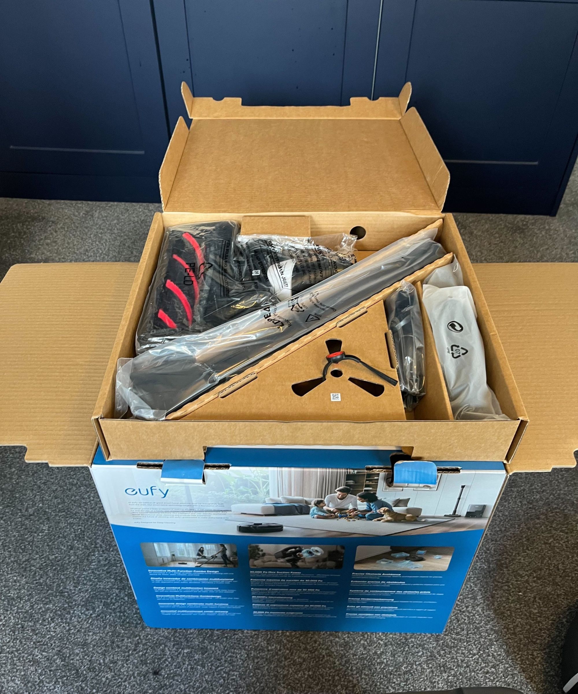 An open blue eufy box on gray carpet with blue closet in the background. All the attachment elements of the vacuum are neatly packaged with the stick, brush head and cleaning arm visible.