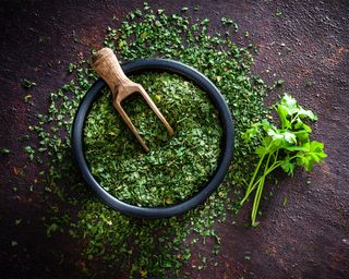 Finely chopped parsley in a small black bowl with wooden server, fresh bunch of parsley on the right, dark textured tabletop