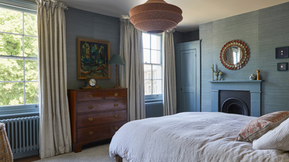 a bedroom with blue grasscloth wallpaper, a wooden dresser upon which rests a clock and a lamp, a hanging pendant lamp, a large bed with white sheets and pillows, and two windows with long curtains and radiators underneath them
