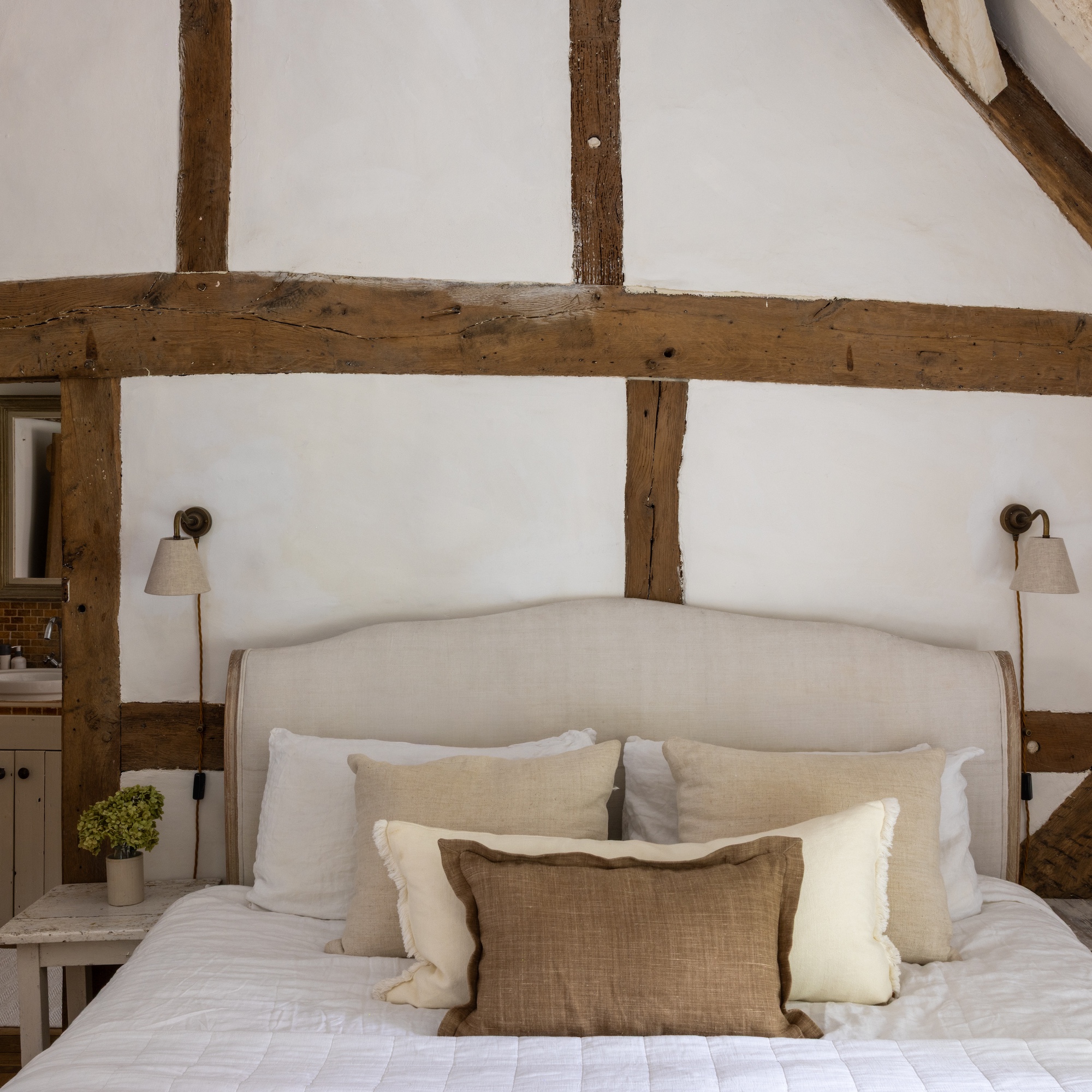 bedroom with old beamed walls and upholstered bed in neutral linen fabrics dressed with linen cushions
