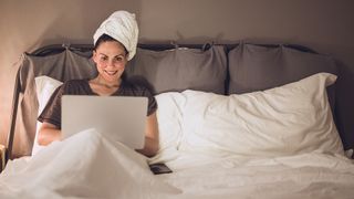 A woman sits on bed using her laptop with her wet hair wrapped in a towel