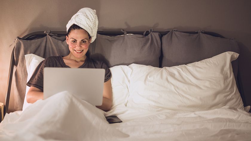 A woman sits on bed using her laptop with her wet hair wrapped in a towel