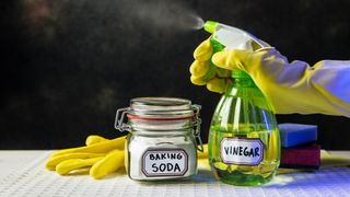 Someone wearing gloves spraying a bottle of white vinegar next to a jar of baking soda